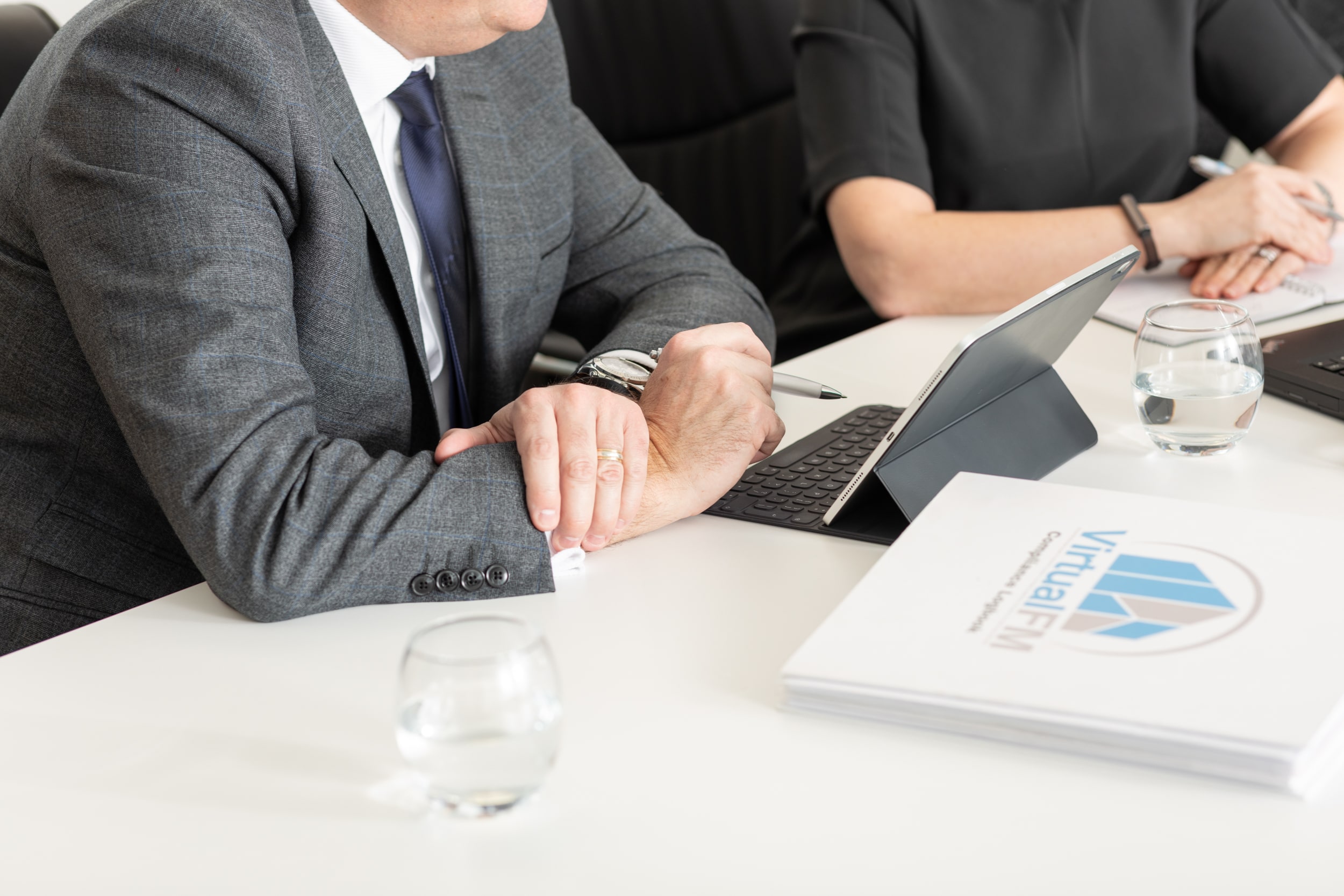 man and woman sitting at desk with hands crossed, tablet on stand on table with logo featuring Virtual FM logo on show